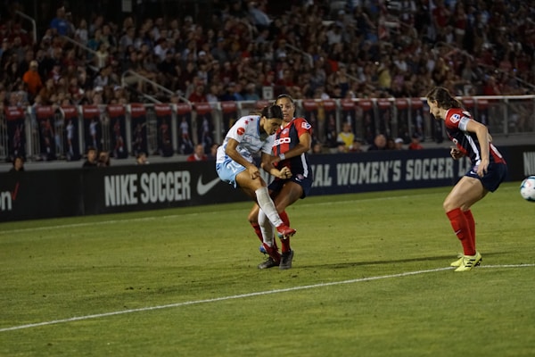 A soccer match is taking place with players in action, one wearing a white and light blue uniform and two others in red and blue uniforms. They are surrounded by a crowded stadium with bright lights and advertisements, indicating possible sponsorships like Nike.