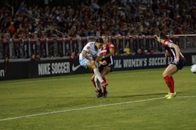 A soccer match is taking place with players in action, one wearing a white and light blue uniform and two others in red and blue uniforms. They are surrounded by a crowded stadium with bright lights and advertisements, indicating possible sponsorships like Nike.