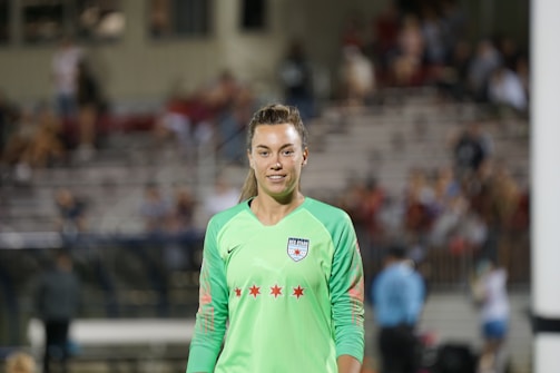 A female soccer player wearing a bright green jersey with a logo and red stars is standing on a soccer field. The background shows blurred spectators seated in a grandstand, suggesting a live match or event.