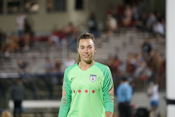 A female soccer player wearing a bright green jersey with a logo and red stars is standing on a soccer field. The background shows blurred spectators seated in a grandstand, suggesting a live match or event.
