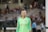 A female soccer player wearing a bright green jersey with a logo and red stars is standing on a soccer field. The background shows blurred spectators seated in a grandstand, suggesting a live match or event.