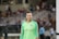 A female soccer player wearing a bright green jersey with a logo and red stars is standing on a soccer field. The background shows blurred spectators seated in a grandstand, suggesting a live match or event.