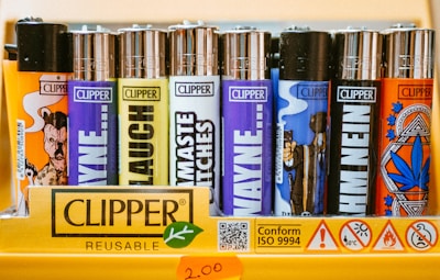 A collection of colorful Clipper lighters is displayed on a shelf. The lighters have various designs and text on them, with each one exhibiting different colors and illustrations. The display also includes branding and safety information printed on the packaging.