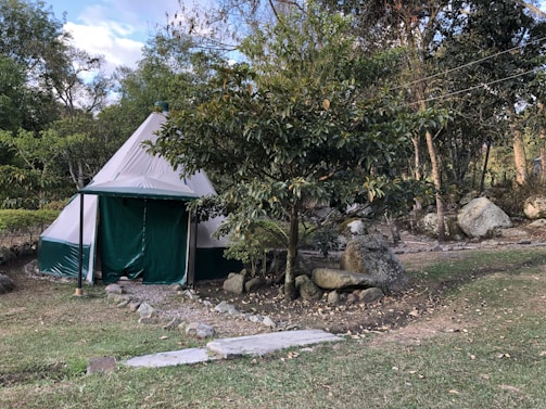 A canvas tent sits in a natural setting surrounded by trees and large rocks. The tent has a conical shape and is primarily white with green accents. Paths lined with stones lead up to the tent, and there is a grassy area in the foreground. The scene suggests a tranquil, outdoor environment.