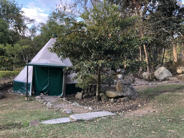 A canvas tent sits in a natural setting surrounded by trees and large rocks. The tent has a conical shape and is primarily white with green accents. Paths lined with stones lead up to the tent, and there is a grassy area in the foreground. The scene suggests a tranquil, outdoor environment.
