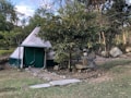 A canvas tent sits in a natural setting surrounded by trees and large rocks. The tent has a conical shape and is primarily white with green accents. Paths lined with stones lead up to the tent, and there is a grassy area in the foreground. The scene suggests a tranquil, outdoor environment.