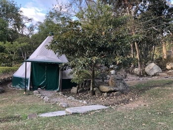 A canvas tent sits in a natural setting surrounded by trees and large rocks. The tent has a conical shape and is primarily white with green accents. Paths lined with stones lead up to the tent, and there is a grassy area in the foreground. The scene suggests a tranquil, outdoor environment.