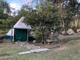 A canvas tent sits in a natural setting surrounded by trees and large rocks. The tent has a conical shape and is primarily white with green accents. Paths lined with stones lead up to the tent, and there is a grassy area in the foreground. The scene suggests a tranquil, outdoor environment.