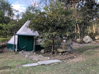 A canvas tent sits in a natural setting surrounded by trees and large rocks. The tent has a conical shape and is primarily white with green accents. Paths lined with stones lead up to the tent, and there is a grassy area in the foreground. The scene suggests a tranquil, outdoor environment.