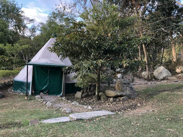 A canvas tent sits in a natural setting surrounded by trees and large rocks. The tent has a conical shape and is primarily white with green accents. Paths lined with stones lead up to the tent, and there is a grassy area in the foreground. The scene suggests a tranquil, outdoor environment.