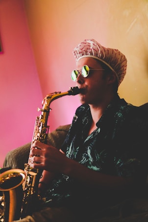 A person wearing a patterned hat and sunglasses is playing a saxophone. The background is illuminated with a warm, colorful gradient. The individual&rsquo;s attire features a dark shirt with a subtle design.