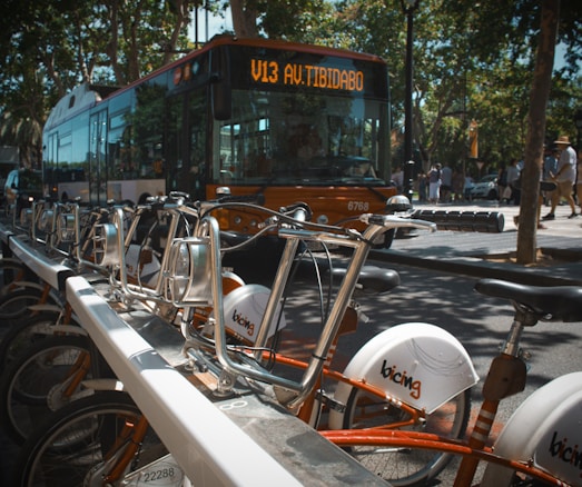 A row of parked bicycles in the foreground, with a public bus displaying the route 'V13 Av. Tibidabo' in the background. The scene takes place on a sunny day, with trees lining the street and people walking on the sidewalk.