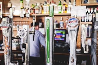 A close-up view of several beer taps in a bar, featuring brands such as Heineken, Coors Light, and Bulmer's. The background shows a variety of bottles and a blurred figure of a person, possibly a bartender, indicating a well-stocked bar environment.
