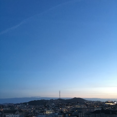 A panoramic view of a city skyline at dusk with scattered city lights beginning to illuminate the scene. A prominent tower stands against the backdrop of a darkening sky, painted with shades of blue and a hint of orange near the horizon.