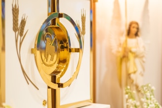 A golden tabernacle with an embossed image of a lamb and cross is displayed in the foreground. Surrounding the tabernacle are decorative engravings of wheat. In the background, a statue of an angel with a staff is partially visible, accompanied by ambient lighting and some white flowers.