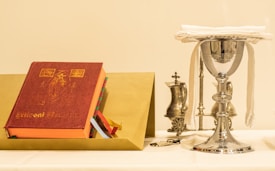 A ceremonial setup includes a large red book with ornate gold lettering and multiple ribbon bookmarks. Beside it, a silver chalice covered with a white cloth sits elegantly, accompanied by two smaller silver vessels and a candlestick in the background. The ensemble is arranged on a white tablecloth.