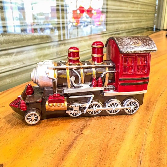 A detailed miniature model of a vintage steam locomotive with vibrant colors, prominently featuring red and black with gold accents. The train is positioned on a wooden surface, with a blurred background showcasing a reflective window.