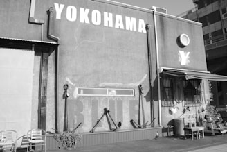 A building's exterior with large, white letters spelling 'YOKOHAMA' on a textured wall. There are metal pipes and a small canopy above some windows on the right. Two chairs, an anchor, and various small items are present against the wall in the foreground.
