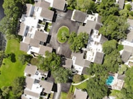 The image is an aerial view of a residential neighborhood featuring multiple houses with brown and gray rooftops. The houses are surrounded by lush green trees and lawns. A cul-de-sac is present in the center with a grassy island. There is a swimming pool on the bottom right corner of the image.