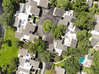 The image is an aerial view of a residential neighborhood featuring multiple houses with brown and gray rooftops. The houses are surrounded by lush green trees and lawns. A cul-de-sac is present in the center with a grassy island. There is a swimming pool on the bottom right corner of the image.