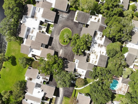 The image is an aerial view of a residential neighborhood featuring multiple houses with brown and gray rooftops. The houses are surrounded by lush green trees and lawns. A cul-de-sac is present in the center with a grassy island. There is a swimming pool on the bottom right corner of the image.