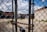 A chain-link fence in the foreground with a heavy chain and padlock. Behind the fence, a gravel-filled lot with sparse grass patches is visible. Graffiti art and a butterfly mural are painted on a brick wall in the background. Overhead utility lines and an overcast sky add an urban atmosphere.