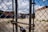 A chain-link fence in the foreground with a heavy chain and padlock. Behind the fence, a gravel-filled lot with sparse grass patches is visible. Graffiti art and a butterfly mural are painted on a brick wall in the background. Overhead utility lines and an overcast sky add an urban atmosphere.