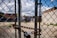 A chain-link fence in the foreground with a heavy chain and padlock. Behind the fence, a gravel-filled lot with sparse grass patches is visible. Graffiti art and a butterfly mural are painted on a brick wall in the background. Overhead utility lines and an overcast sky add an urban atmosphere.