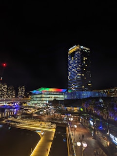 A nighttime urban scene featuring a brightly lit cityscape. A tall building with the name 'Sofitel' on top is prominent, surrounded by other structures illuminated by colorful lights. A pedestrian walkway with street lamps leads towards the buildings, and a glass-fronted structure displays a spectrum of colors on its roof. The area around is bustling with people, giving the environment a lively vibe.