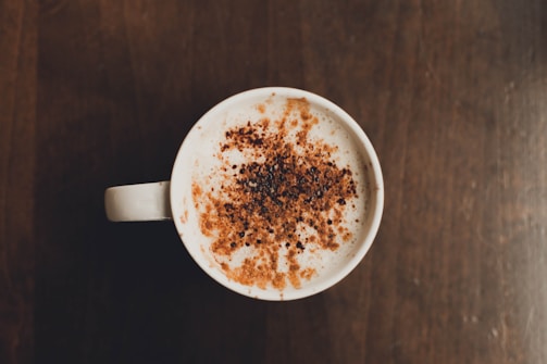 A top view of a cup filled with frothy coffee or latte, topped with a generous sprinkle of brown powder, possibly cinnamon or cocoa. The cup is placed on a dark wooden surface.