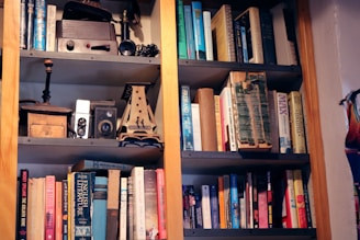A bookshelf filled with an assortment of books and various vintage items. The books range in size and color, and are arranged on multiple shelves. On the top shelf, there are old cameras, a wooden box, and other vintage artifacts.