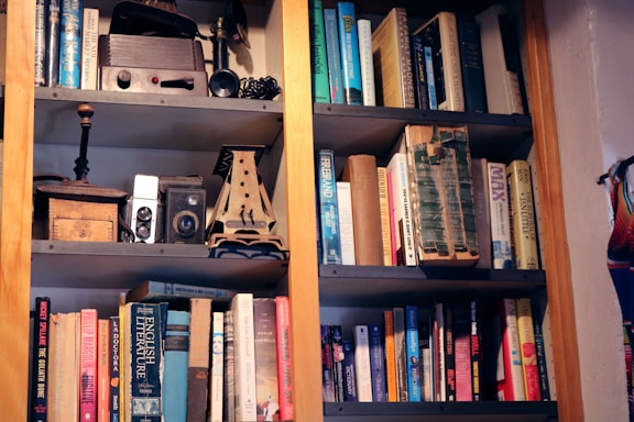 A bookshelf filled with an assortment of books and various vintage items. The books range in size and color, and are arranged on multiple shelves. On the top shelf, there are old cameras, a wooden box, and other vintage artifacts.
