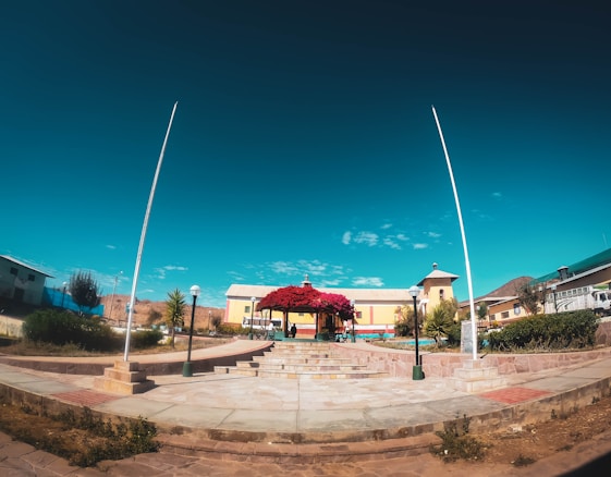A vibrant plaza with a central gazebo covered in bright red flowers, surrounded by well-maintained gardens and a few small buildings in the background. The sky above is clear and blue, adding a crisp contrast to the earthy tones of the plaza.