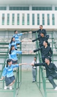 A group of young people stands on a set of tiered metal structures, with some wearing blue traditional attire and others in black. They are positioned in such a way that their hands are reaching out towards each other. The background features a modern building with several windows.