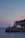 A scenic coastal landscape featuring a striped black and white lighthouse situated on a rocky cliff. The lighthouse is accompanied by a few adjacent buildings, set against the backdrop of a serene sea and a pastel-colored sky at sunset.