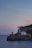A scenic coastal landscape featuring a striped black and white lighthouse situated on a rocky cliff. The lighthouse is accompanied by a few adjacent buildings, set against the backdrop of a serene sea and a pastel-colored sky at sunset.