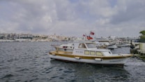 Several boats are docked at a marina with one prominently displaying the Turkish flag. The water is calm and the skyline in the background shows a densely built urban area under a cloudy sky.