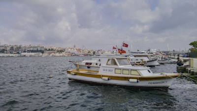 Several boats are docked at a marina with one prominently displaying the Turkish flag. The water is calm and the skyline in the background shows a densely built urban area under a cloudy sky.