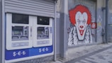 A pair of automated teller machines (ATMs) are mounted on an outdoor wall. Next to them, there is a large graffiti mural of a clown with red hair and a big smile painted on the gray wall. The ground is paved with stone tiles.