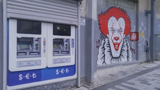 A pair of automated teller machines (ATMs) are mounted on an outdoor wall. Next to them, there is a large graffiti mural of a clown with red hair and a big smile painted on the gray wall. The ground is paved with stone tiles.