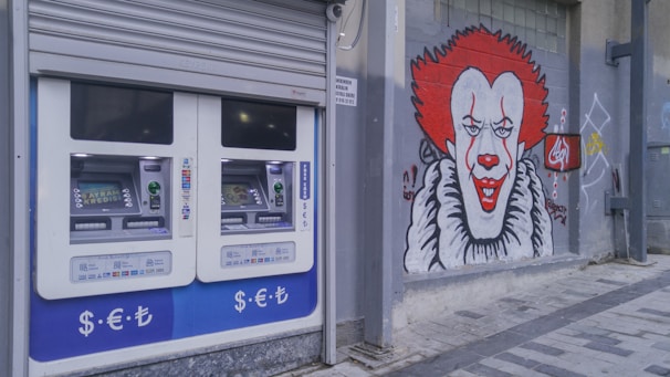A pair of automated teller machines (ATMs) are mounted on an outdoor wall. Next to them, there is a large graffiti mural of a clown with red hair and a big smile painted on the gray wall. The ground is paved with stone tiles.