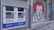 A pair of automated teller machines (ATMs) are mounted on an outdoor wall. Next to them, there is a large graffiti mural of a clown with red hair and a big smile painted on the gray wall. The ground is paved with stone tiles.