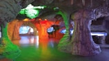 A cave interior featuring multiple stalactites and stalagmites with colorful artificial lighting in green, orange, and purple hues, creating a mystical atmosphere. The cave walls and formations have an undulating texture, and there are passageways leading deeper into the cave.