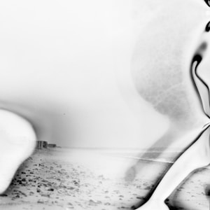 A black and white abstract image combining elements of a beach and a distorted figure. The shoreline is visible with scattered rocks, and the image has a wavy effect that gives an impression of movement or distortion.