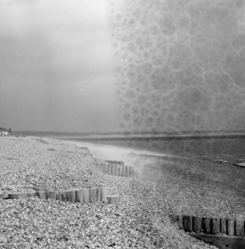 A monochrome beach scene with a pebble-strewn shore and rows of wooden groynes reaching into the water. The ocean appears calm under a cloudy sky, and there is a textured overlay on the right side of the image, creating a mottled effect.