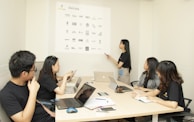 A group of five individuals are in a conference room engaging in a meeting. Four people are seated at a table with laptops and notebooks in front of them, while one person is standing and pointing at a projection on the wall displaying various company logos.