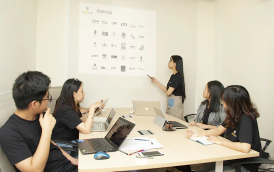 A group of five individuals are in a conference room engaging in a meeting. Four people are seated at a table with laptops and notebooks in front of them, while one person is standing and pointing at a projection on the wall displaying various company logos.