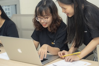 Two people are engaged in collaborative work at a desk, both wearing black t-shirts and focusing on a laptop. One person is seated and typing on the laptop, while the other is leaning over, looking at the screen and holding a pencil. Papers are scattered on the desk.