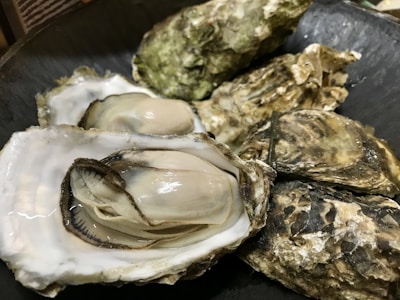 A collection of fresh oysters, some of which are open, revealing their plump meat inside their textured, rough shells. The shells have a variety of shades, from brown to green, and the oysters are resting on a dark surface.