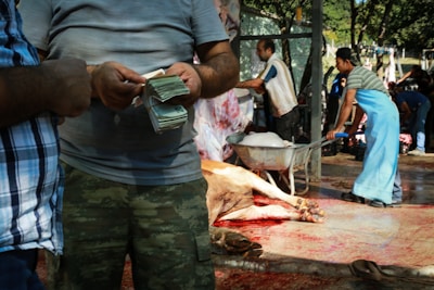 A scene at a slaughterhouse where several people are involved in different tasks. One person is counting a stack of money while another pushes a wheelbarrow filled with meat. In the background, people are busy with the butchering process. The ground is stained with blood, and a cow carcass is visible.